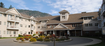 A pink beige three story building with a brown roof and garden in front and mountains and blue sky in back