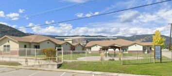 A wide low-slung building with beige stucco and a brown roof with green grass and a parking lot in front with a blue sky background