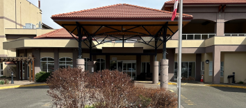 The front of a building with an orange terracotta roof, large pillars, a garden and Canadian flag, and a roundabout driveway in front