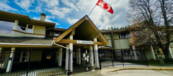 A large facility with a sign featuring the Interior Health logo and the words Bastion Place. A large canadian flag hangs from a slanted pole on the roof attached to the front of the building.