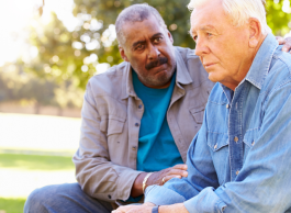 A man sits next to another man on a bench and comforts him.