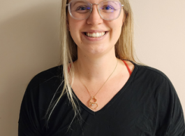 Woman with blonde hair and glasses wearing black top standing against beige wall.