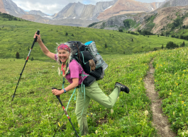 Woman with hiking poles and backpack taking long strides on grass with mountains in the background.