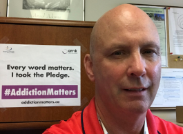 A person stands next to a sign that says “Every word matters. I took the pledge. #AddictionMatters addictionmatters.ca”