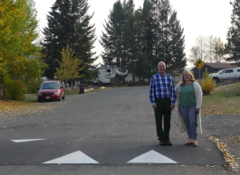 A bald man with a blue plaid shirt and black pants standing to the left of a woman with long blonde hair, beige cardigan, green shirt and blue plaid pants. Both are standing on a speed bump on a residential street.