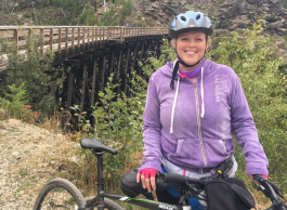 Smiling woman cyclist in purple top and bike helmet, posts by wooden bridge.