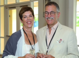 A woman with short brown hair standing on the left with a white dress and white and blue shawl, and a man with grey and white hair and moustache standing on the right with a light pink dress shirt and white suit jacket, with black-framed glasses, with French doors and a wall of windows behind them.