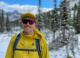 A man in a yellow jacket and ball cap, wearing a blue backpack, standing in front of snowy trees.