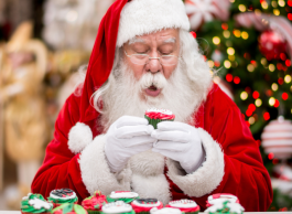 A person dressed up as Santa Claus holds up a piece of Christmas baking, with a table full of Christmas baking in front of them.