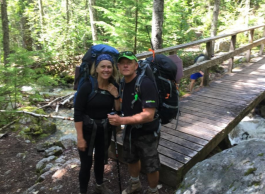 A woman on the left wearing a blue bandana on head, and black clothes, with a large blue backpack, and a man on the right wearing a black shirt and camo shorts, wearing a black backpack, standing in front of a brown bridge over a creek, in a forest.