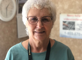 Woman with white hair, glasses, in green top with lanyard. There is a clock and posters on wall behind her.