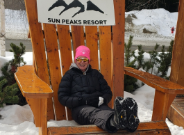 A woman in winter gear sits outside on a big wooden chair with a black Sun Peaks Resort logo on it.