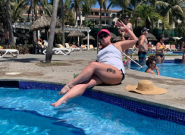 A woman wearing a pink ball cap and white camisole shirt with a tattoo on her upper left thigh, sitting at a pool with palm trees in the background and a straw hat sitting next to her on the pool deck