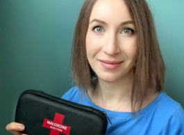 A woman with short brown hair wearing a blue shirt holds up a black case that says Naloxone Injection on it.