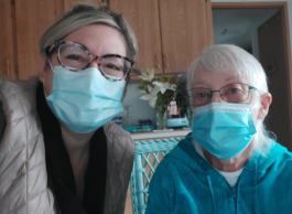 A middle aged and elderly person, both wearing masks, take a selfie inside.