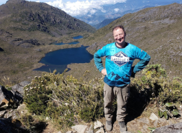 Man smiling with hands on hips, wearing a blue sweatshirt and khaki pants with mountains and lake background.