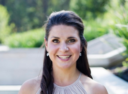 A smiling woman with earrings standing outside. The background is blurred.