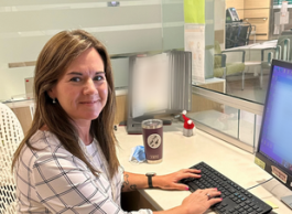 A smiling woman with brown hair and a white checkered shirt poses for a picture while sitting down and typing on a computer.