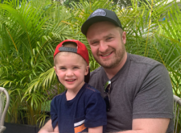 A child, left, sits on an adult’s lap, both smiling, with greenery in the background.