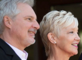 Four people standing side by side: a man with grey hair, a woman with short blonde hair and a pink shirt, a man with brown hair, a beard and wearing a suit, and a woman with long, blonde hair.