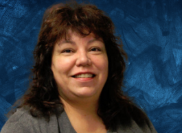 A smiling woman with brown hair and wearing a grey shirt poses for a picture in front of a blue background.