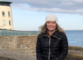 A woman wearing a jacket and a toque stands on a boardwalk with water and a lighthouse behind her.