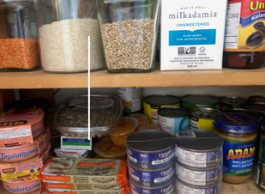 A pantry shelf with beans, rice and lentils in glass jars, macadamia beverage, olives in a jar, cans of fish and sardines, and peanut butter