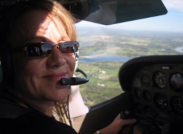 A woman wearing a pilot’s headset is sitting in the cockpit of an aeroplane with the Okanagan landscape visible through the windscreen in the background.