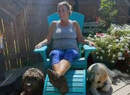 A woman in a tank top and leggings sits on a large green chair in the sunshine, with various potted flowers in the background to her right