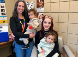 A woman wearing jeans, a black sweater and a lanyard that says Interior Health smiles while holding a toddler. Beside her a younger woman with red hair wearing all black sits in a chair holding an identical toddler.