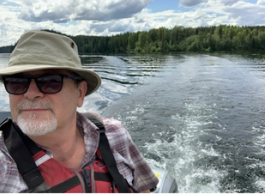 A bearded man wearing a beige hat and sunglasses rides in a boat on a lake.