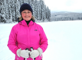 Smiling woman wearing pink jacket, white gloves, wool hat, standing on snow with snow-covered trees and mountains in background