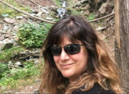 A woman with shoulder-length brown hair and dark sunglasses smiles in front of a creek surrounded by leaves, trees, and rocks.