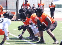 Football players dressed in orange face each other wearing black helmets. One holds a football to the ground. 