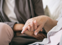 A chest and shoulders picture of a person in a taupe jacket and cream coloured shirt holds the hand of an elderly person wearing a white robe