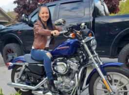 A person in a brown leather jacket and ripped jeans sitting on a blue Harley-Davidson motorcycle in front of a black pick-up truck