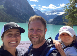 Three people, a mom, dad and baby in a carrier, stand in front of an emerald green lake surrounded by mountains, trees and a blue sky with fluffy clouds