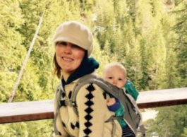 A woman with a cream sweater and white hat wears a backpack that holds a baby. She smiles as she stands in front of trees and water. 