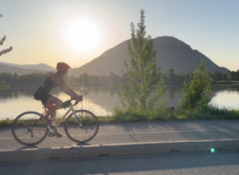 A person on a road bike rides on a path at sunrise next to a lake and mountain