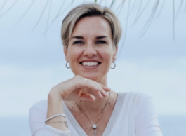 Woman with short blonde hair smiling, with her chin resting on her hand, wearing white, and a white background.