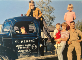 A family of six people, two adults and four children, stand next to, on, and in an old blue truck