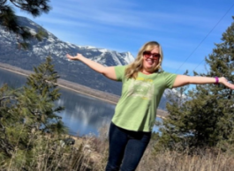 Woman with blonde hair wears sunglasses and smiles as she holds her arms outstretched in front of a background of mountains and a lake