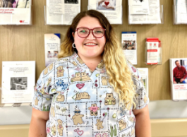 Smiling woman with long hair and glasses, wearing scrubs, in front of wall with information pamphlets