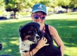 A woman wearing a blue baseball cap and sunglasses is smiling as she holds her dog