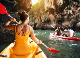 Three people kayak in water between two cliffs with overhanging leaves.