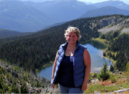 A woman poses for a picture on a tall hill with mountains, trees and water in the background.