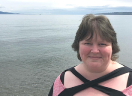 Smiling woman with short hair standing in front of lake with mountains and cloudy sky in the background.