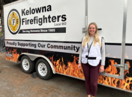 A woman with long blonde hair wearing a white safety vest stands in front of a white Kelowna Firefighters truck that says proudly supporting our community