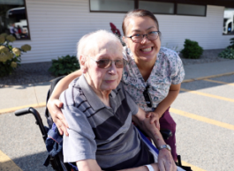 A person in medical clothing puts their arms around a senior in a wheelchair as they pose for a picture in a parking lot outside a building.