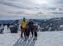 A family of four, two adults and two children, wearing ski equipment and helmets pose on a ski run with mountains and clouds in the background